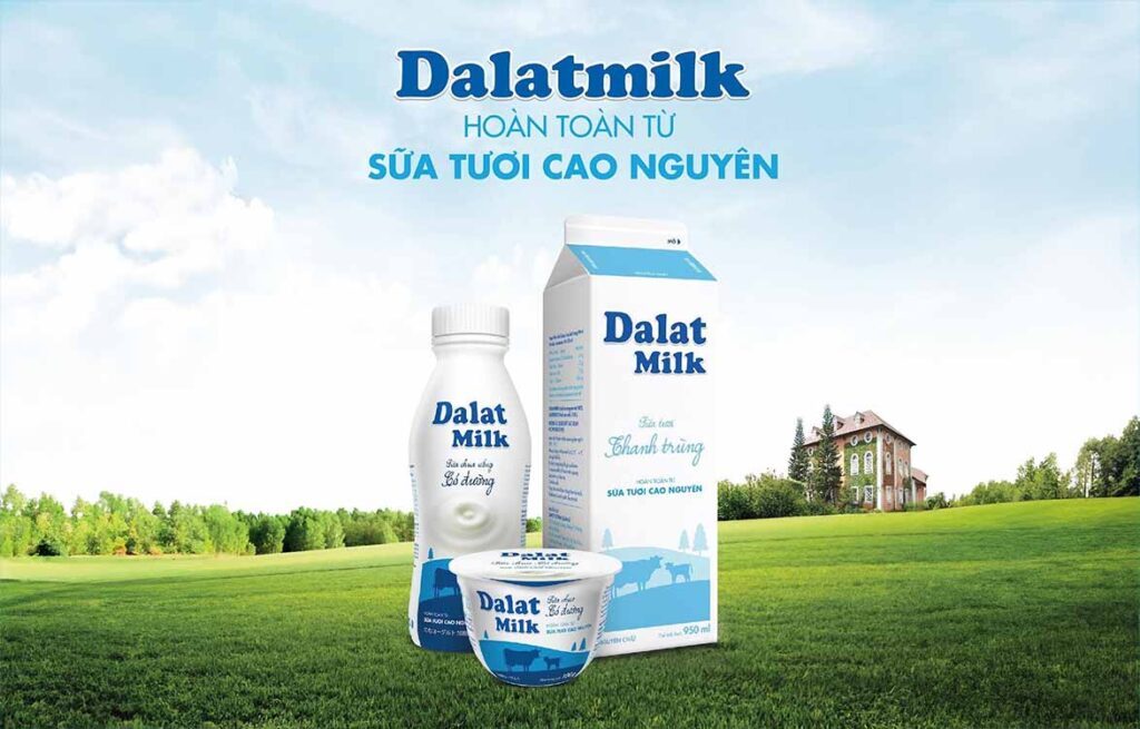 3 sản phẩm sữa Dalat milk bao gồm: sữa chua uống dalat milk có đường 500ml, sữa chua ăn dalat milk có đường 100g và sữa thanh trùng dalat milk không đường 950ml đang ở trên cánh đồng cỏ màu xanh, phía sau xa xa là nhà máy sản xuất của dalat milk